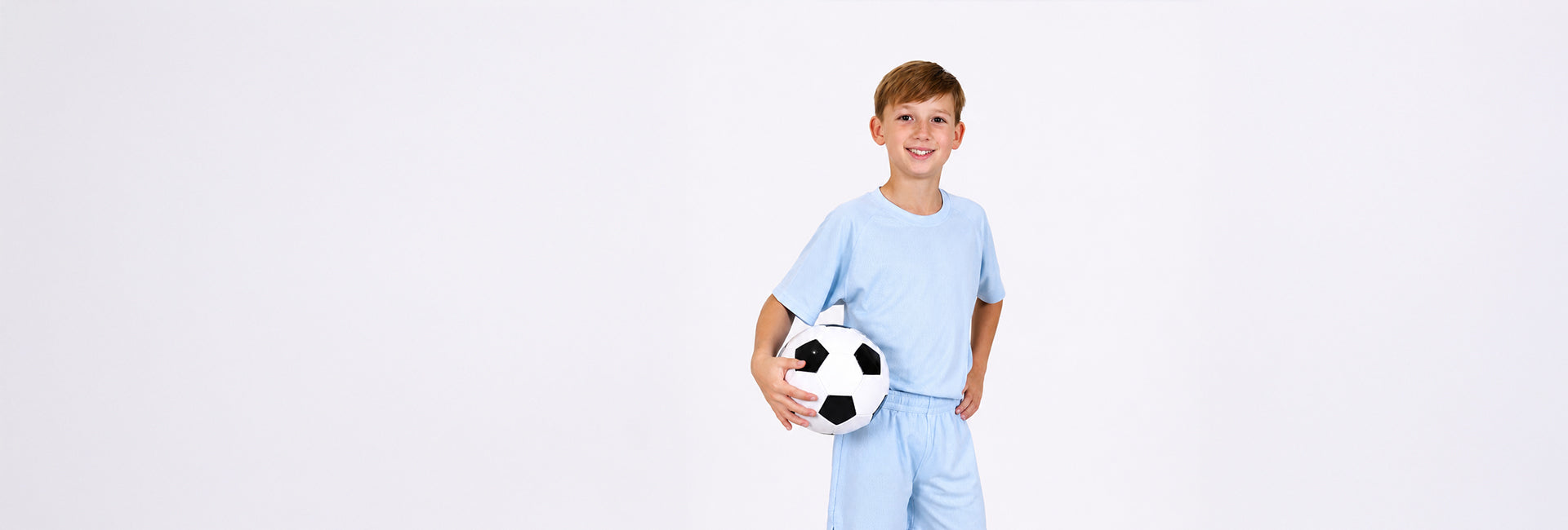 Boy aged ten holding a football under his arm with a regenerative cotton hoody over his shoulders and wearing sky blue sports shorts and sports t-shirt made from toxin free, natural materials 