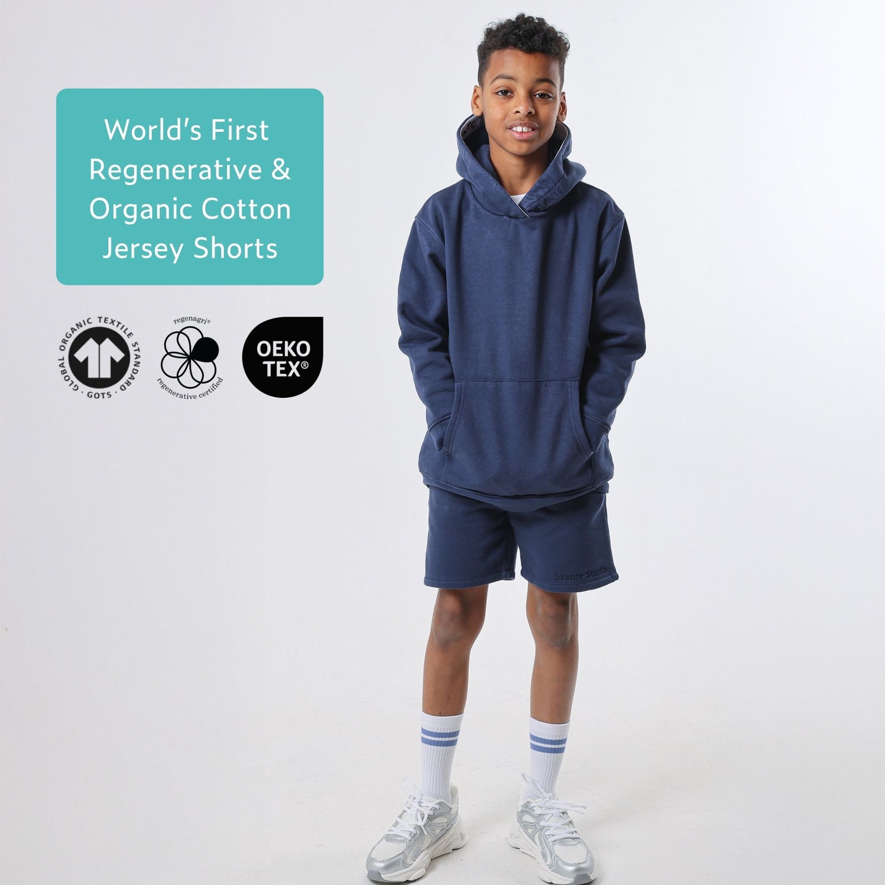 Boy wearing a navy blue organic cotton hoodie and  blue organic cotton jersey shorts with certification logos on a white background. #Peacoat-Blue