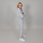Blonde kid standing side on with arms folded, wearing a grey organic joggers and hoodie  with white sneakers in a light studio