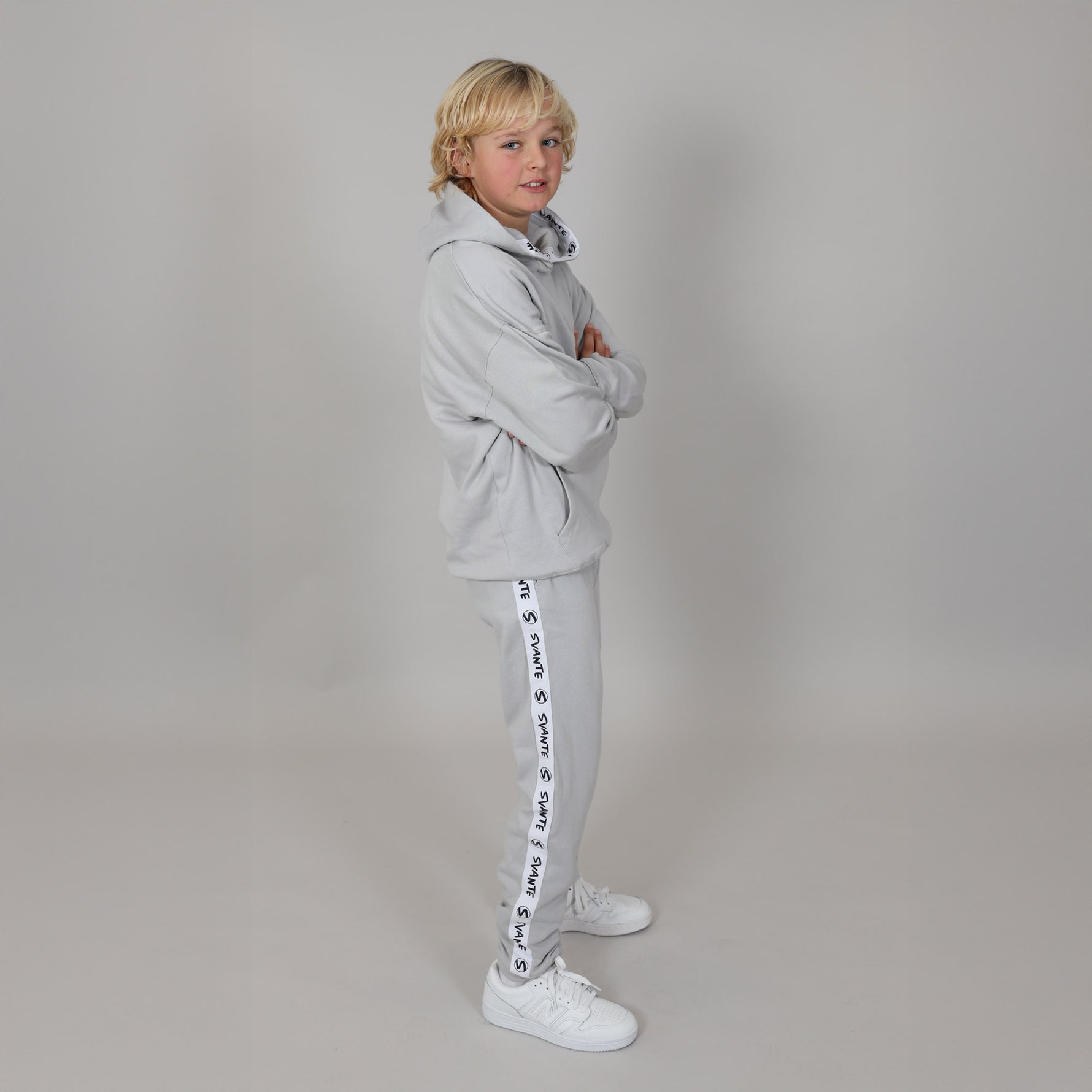 Blonde kid standing side on with arms folded, wearing a grey organic joggers and hoodie  with white sneakers in a light studio