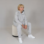 Child wearing a gray organic tracksuit with white sneakers sitting on a beige stool against a plain background