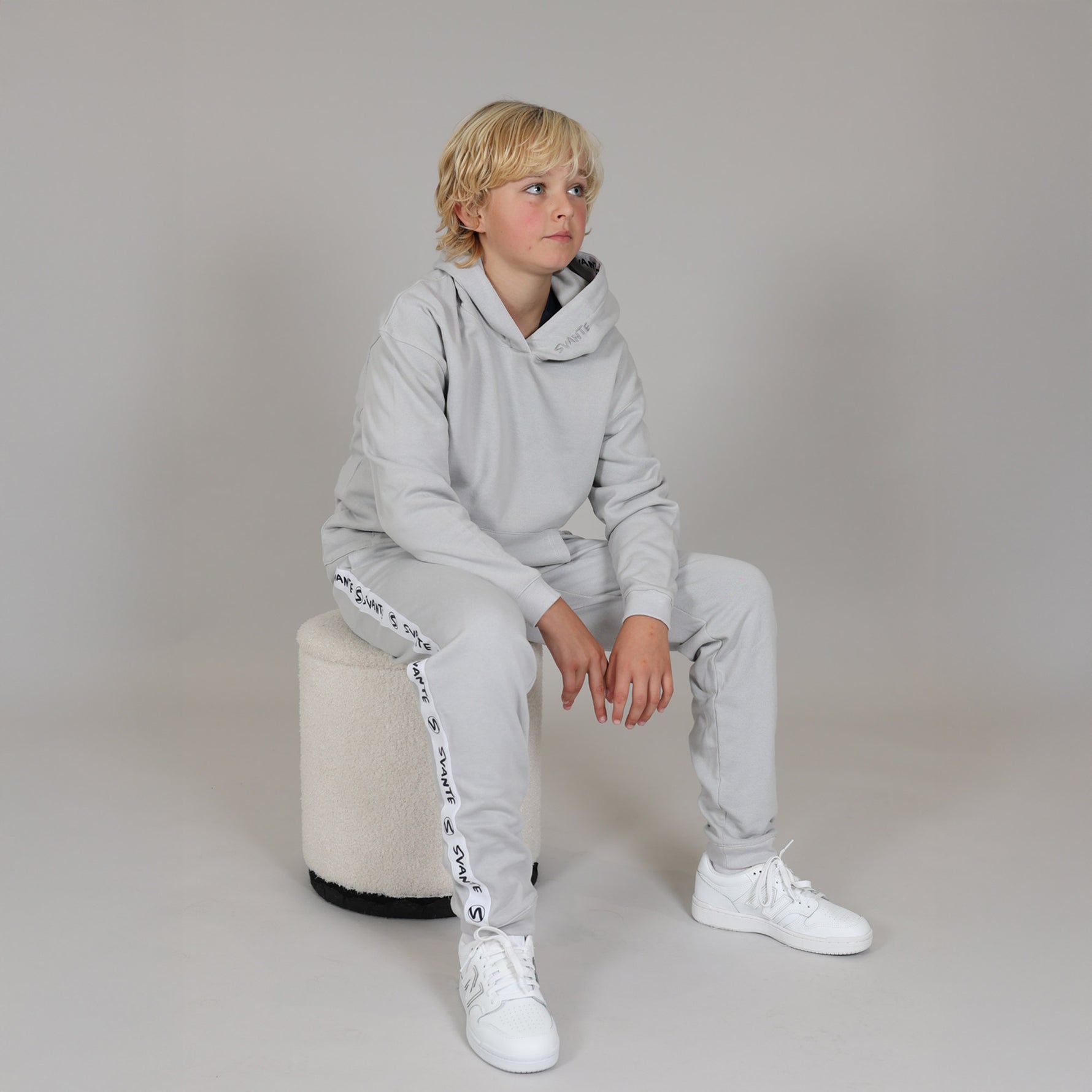 Child wearing a gray organic tracksuit with white sneakers sitting on a beige stool against a plain background