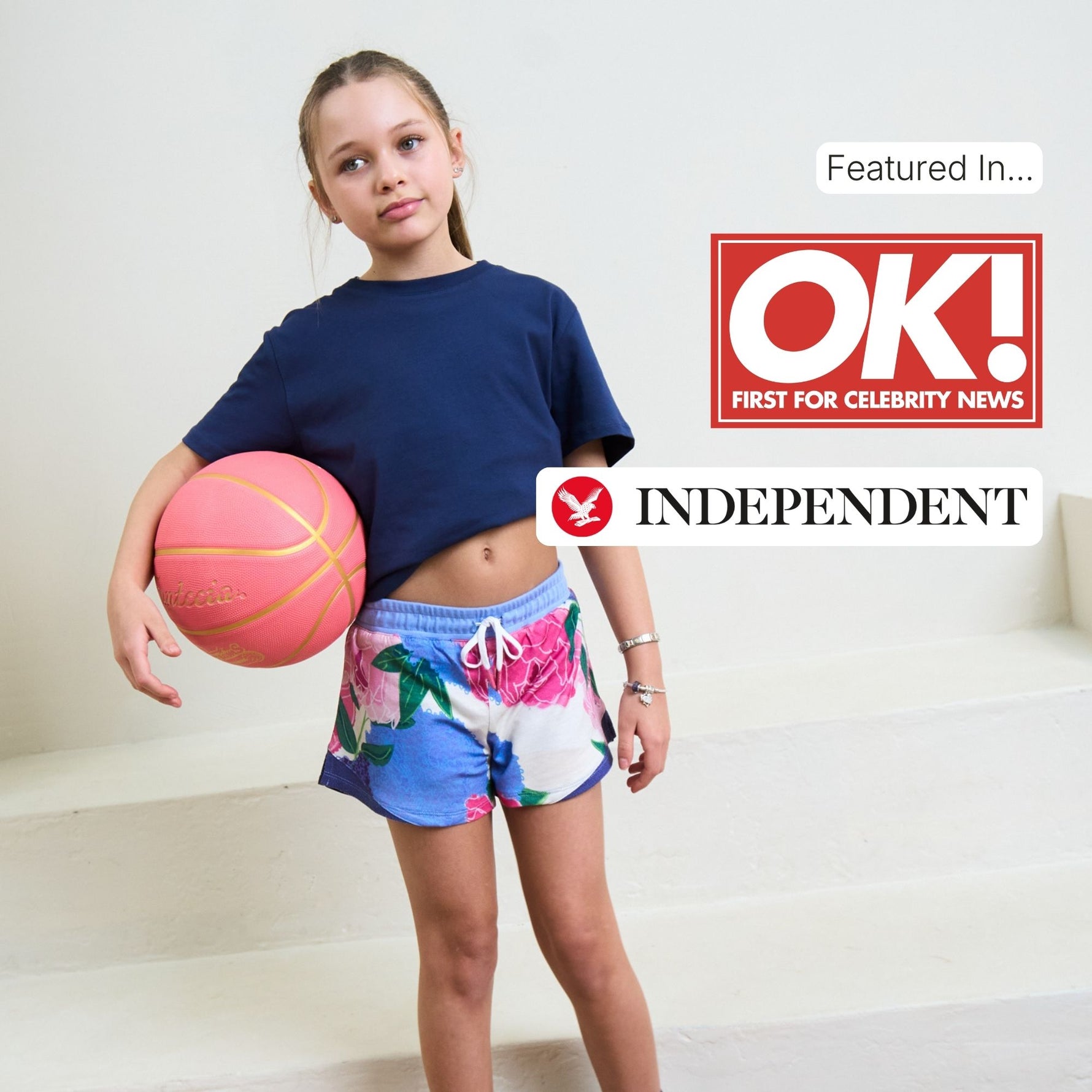 Girl holding a pink basketball with 'OK!' and 'Independent' logos above her, she is wearing a navy blue organic cotton t shirt and plastic free blue sports shorts. 