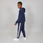 Kid standing side on wearing a Svante navy tracksuit with Svante logo in white text running down the leg, on a plain background. 