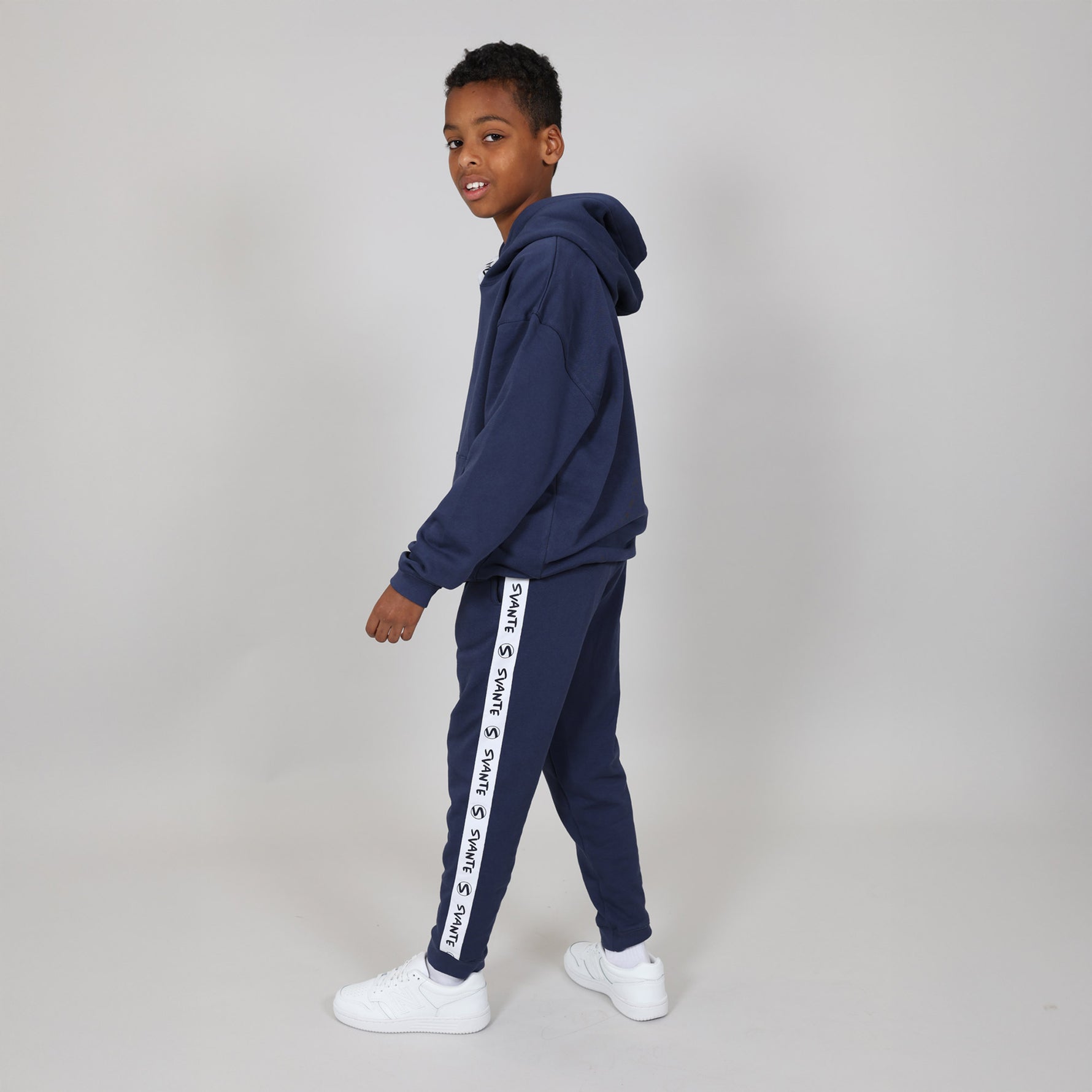 Kid standing side on wearing a Svante navy tracksuit with Svante logo in white text running down the leg, on a plain background. 