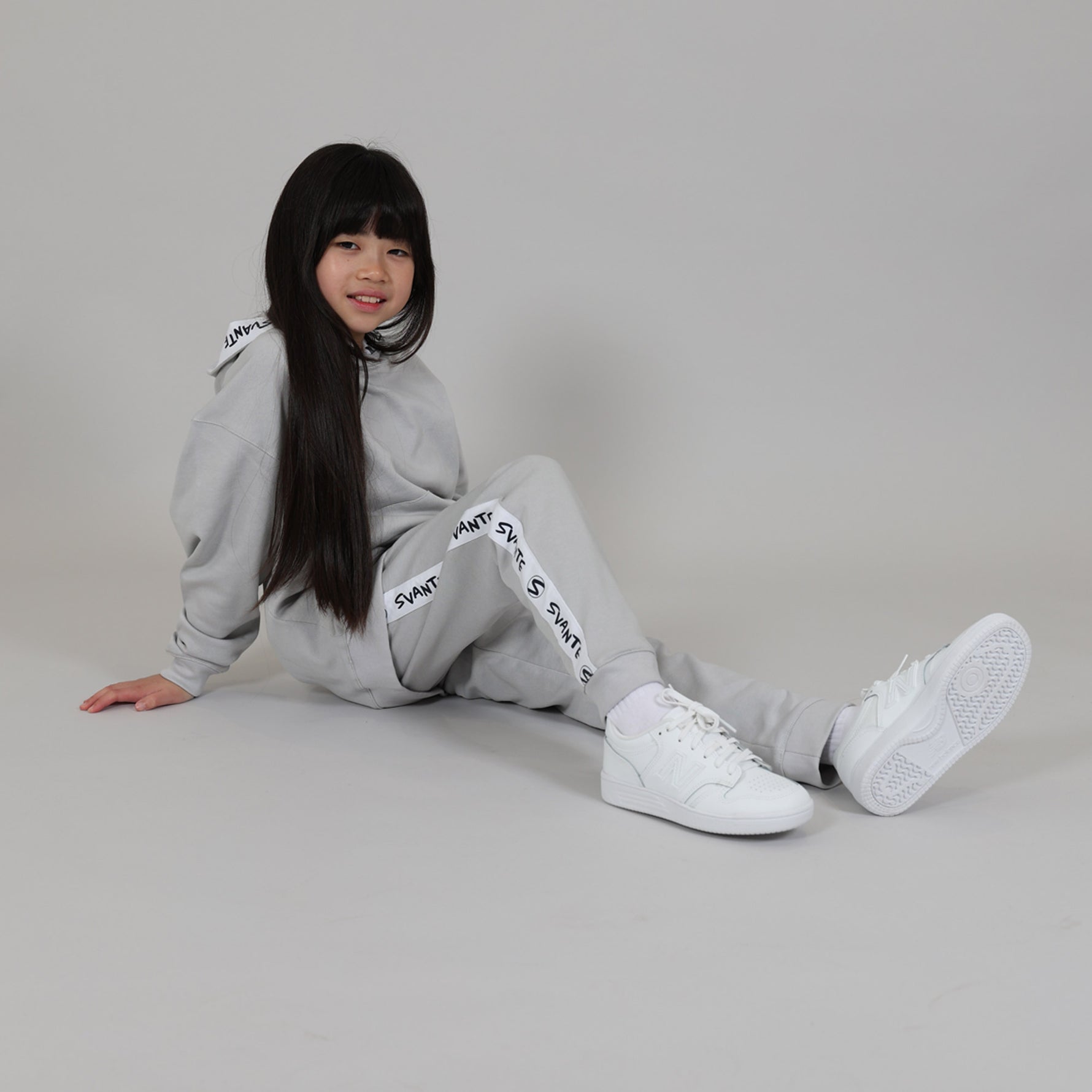 Child sitting on the floor with knee up wearing a grey tracksuit with white sneakers in a studio