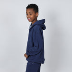Boy aged 11 standing side on wearing a navy blue organic hoodie on a plain background. 