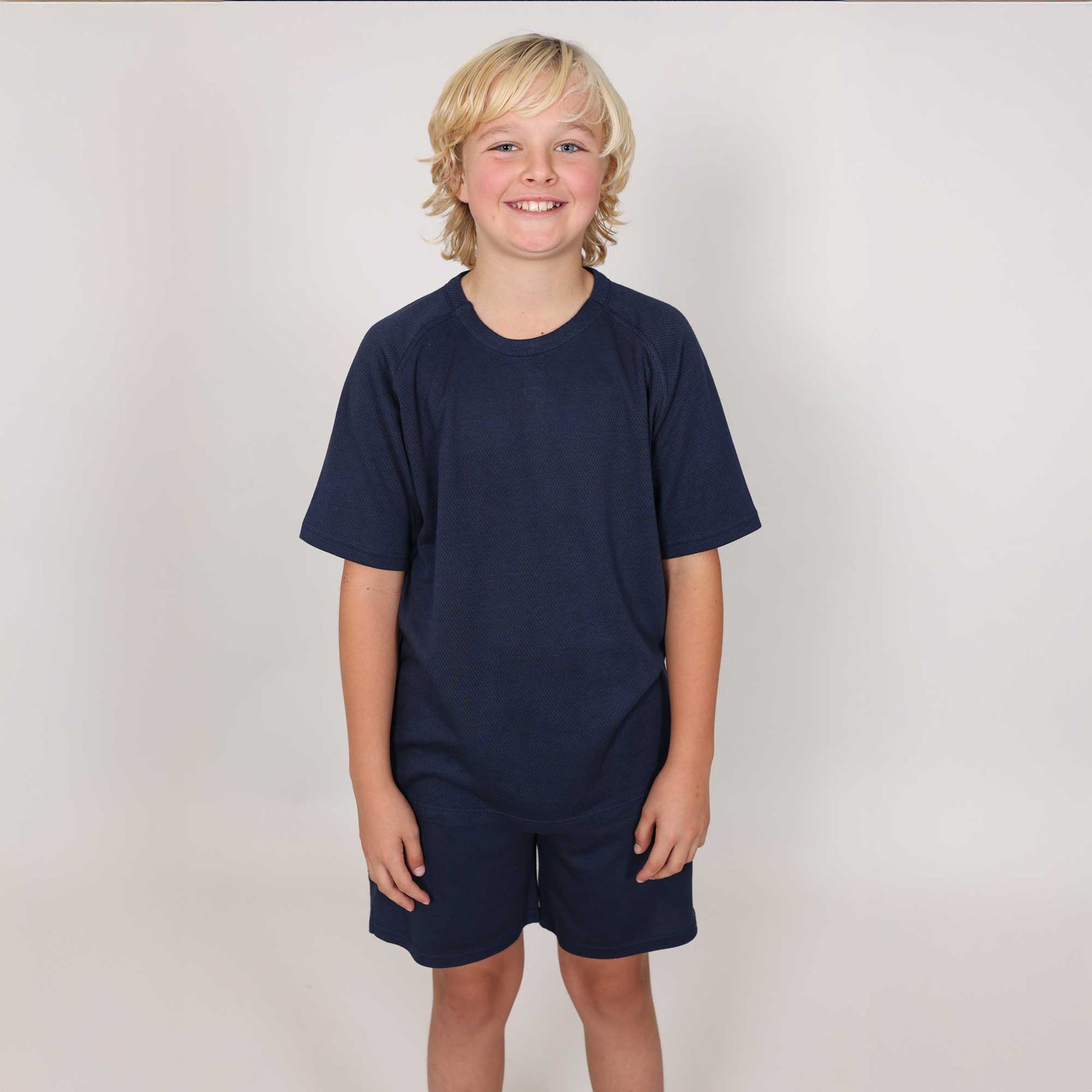 Young boy with big smile wearing a navy blue sports t-shirt and shorts against a plain grey background