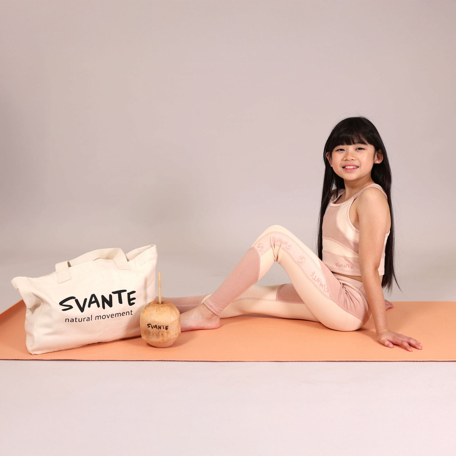 Young girl in a pink sports outfit sitting on an orange yoga mat with a 'SVANTE' organic cotton tote bag and coconut drink beside her.
