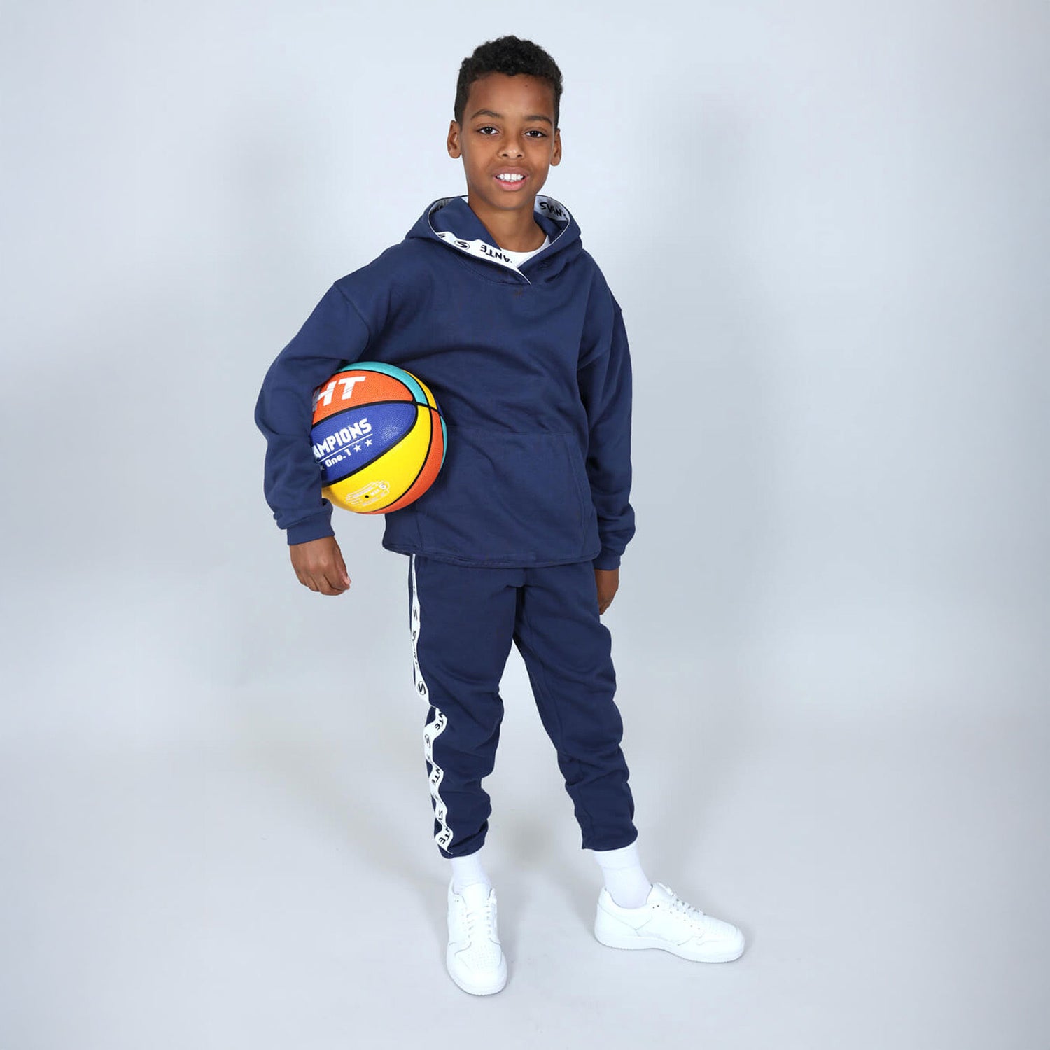 Boy holding a basketball wearing a Svante regenerative cotton navy blue tracksuit with white trainers on a grey and white background. 