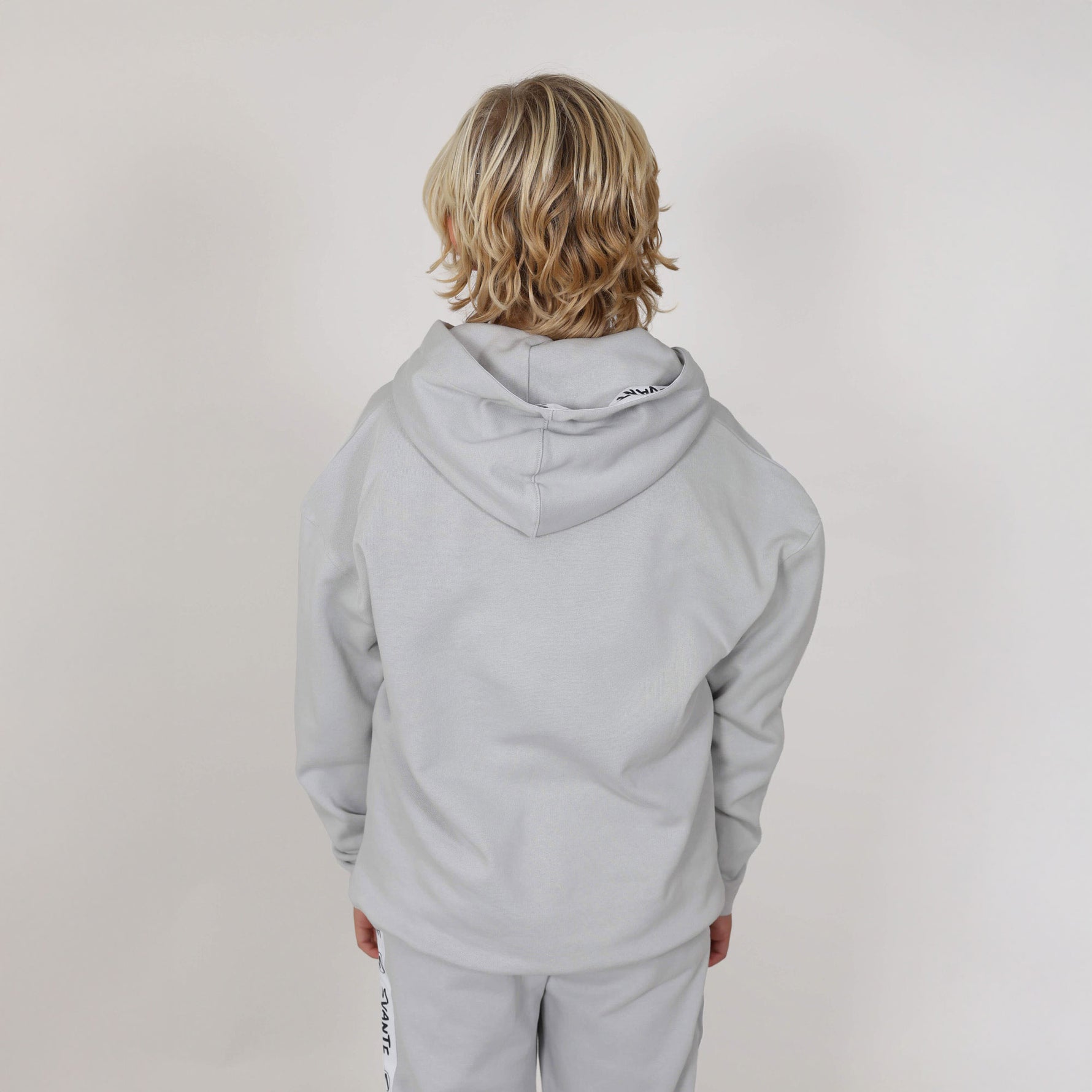 Boy with blonde hair with back to camera showing the reverse side of a grey regenerative hoodie and joggers in professional studio. 
