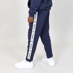 Kid standing side on and wearing white trainers and regenerative cotton navy joggers with white 'Svante' logo down the leg on a grey background. 