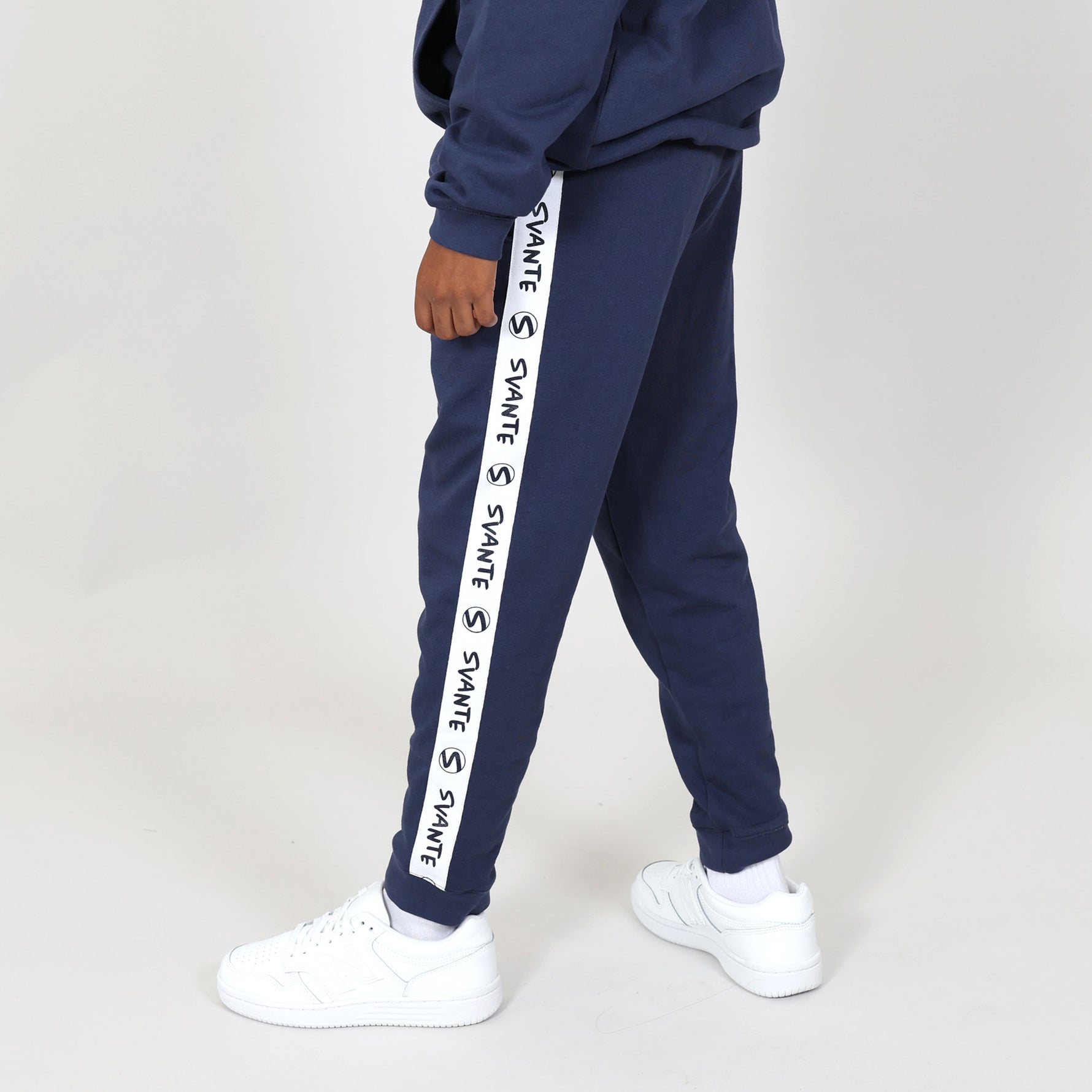 Kid standing side on and wearing white trainers and regenerative cotton navy joggers with white 'Svante' logo down the leg on a grey background. #Peacoat-Blue