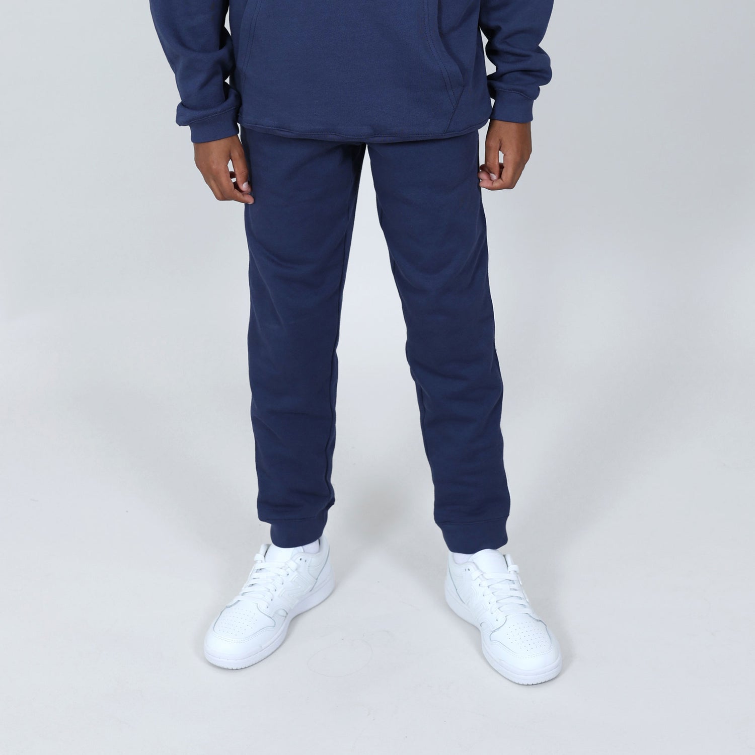 Boy wearing Svante regenerative cotton navy joggers with organic cotton stitching, wearing white sneakers on plain grey background. 