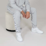Boy sat on a low stool wearing a grey organic cotton tracksuit with white sneakers on a neutral background. 