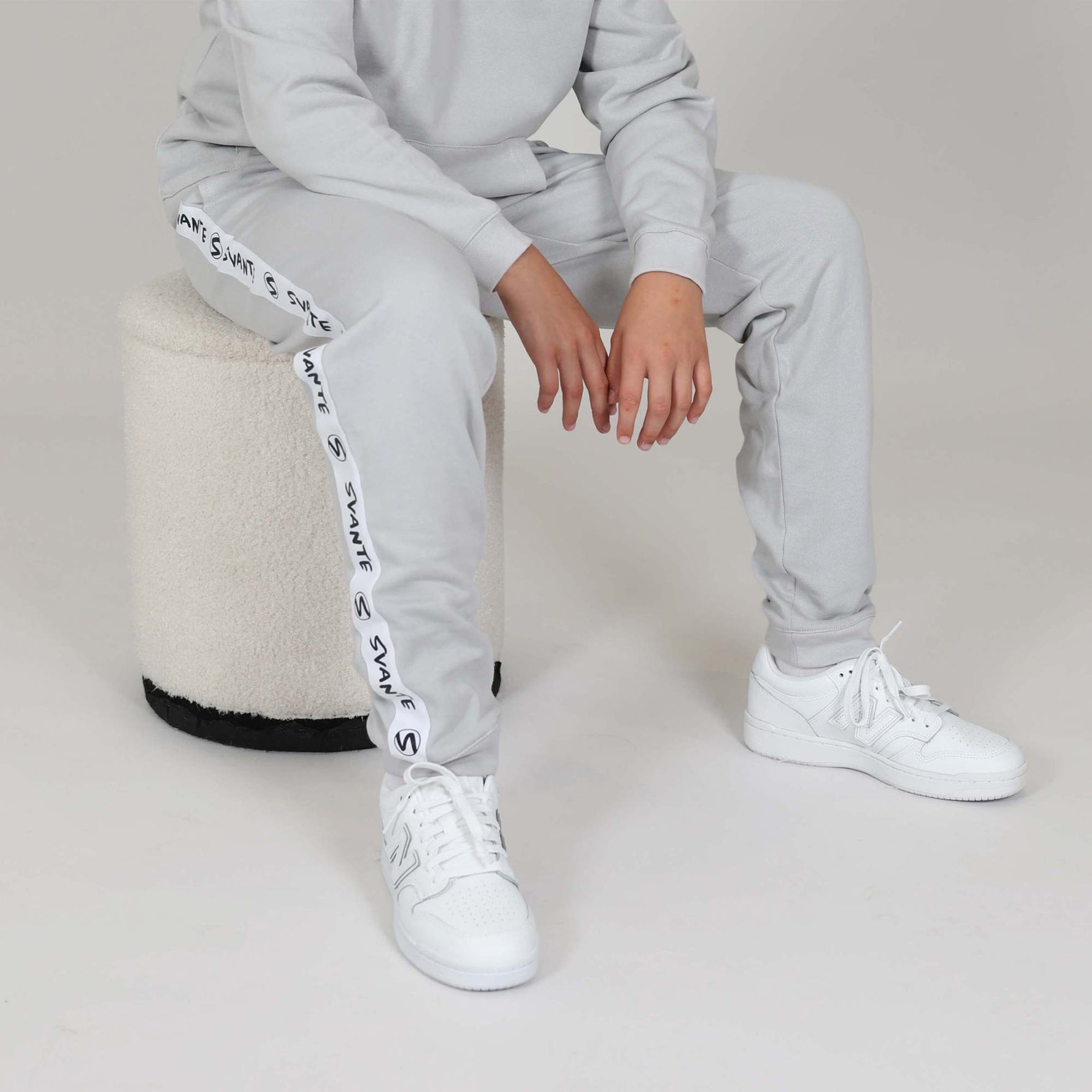 Boy sat on a low stool wearing a grey organic cotton tracksuit with white sneakers on a neutral background. 