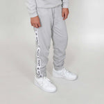 Low angle image of a boy wearing light regenerative cotton grey jogger pants with white Svante tape down the leg and white sneakers on a calm background. 