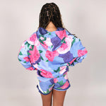 Teenage girl with back to camera wearing a colourful floral hoodie and shorts set on a plain background. 
