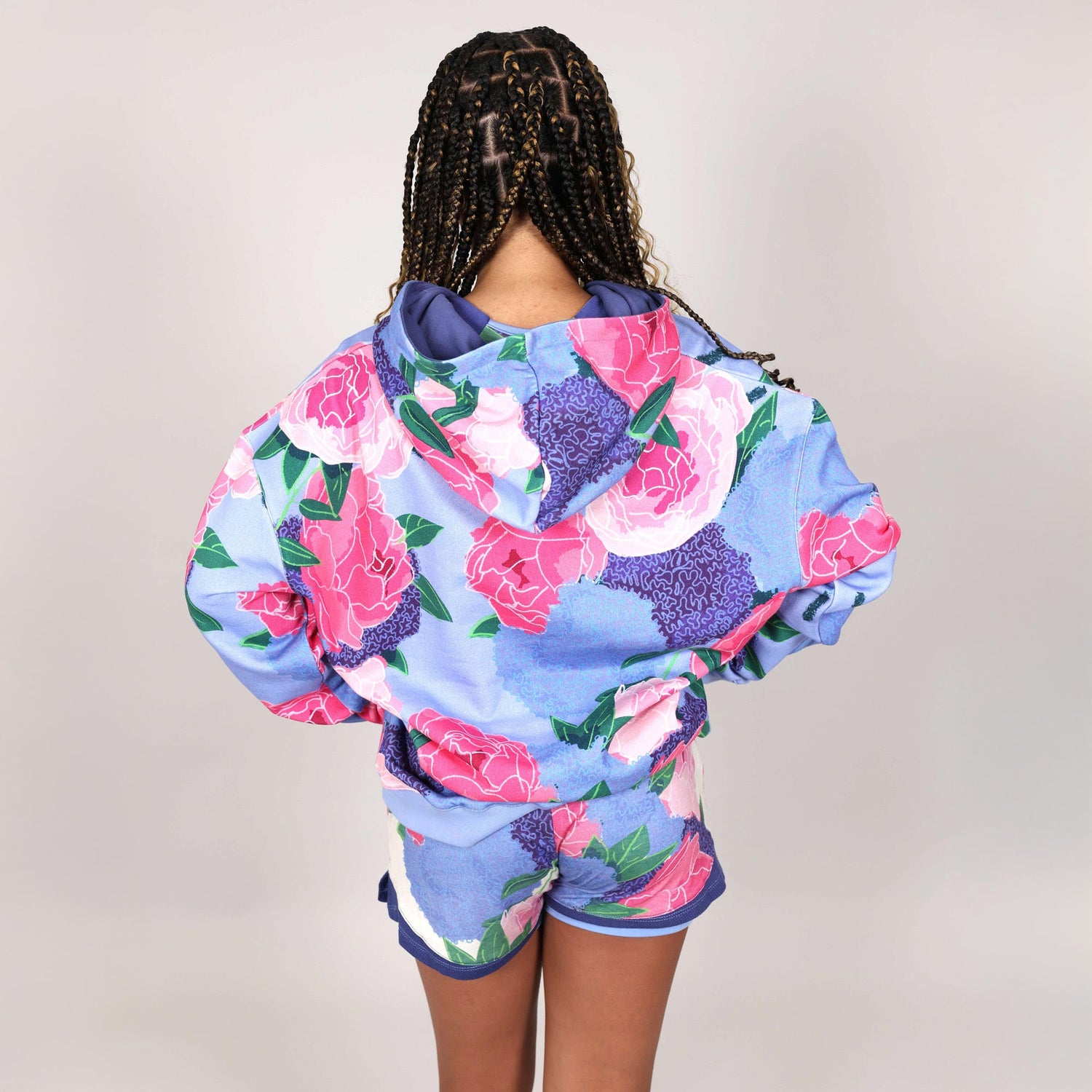 Teenage girl with back to camera wearing a colourful floral hoodie and shorts set on a plain background. 