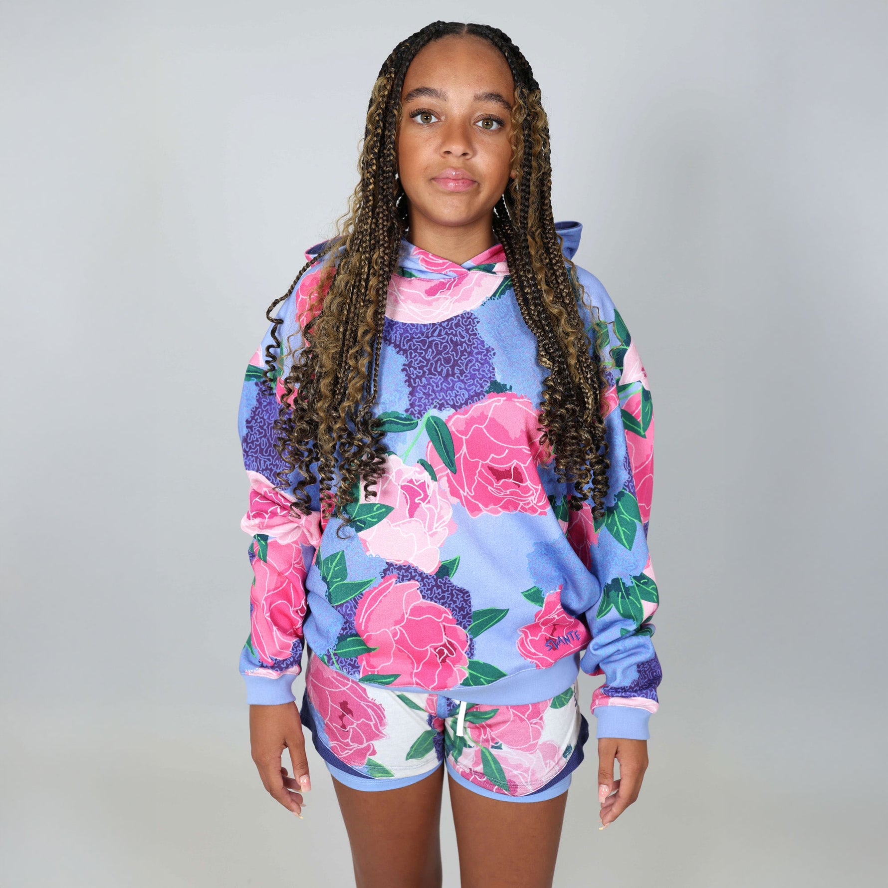 Teenage girl wearing a colourful floral hoodie and shorts on a grey background. 