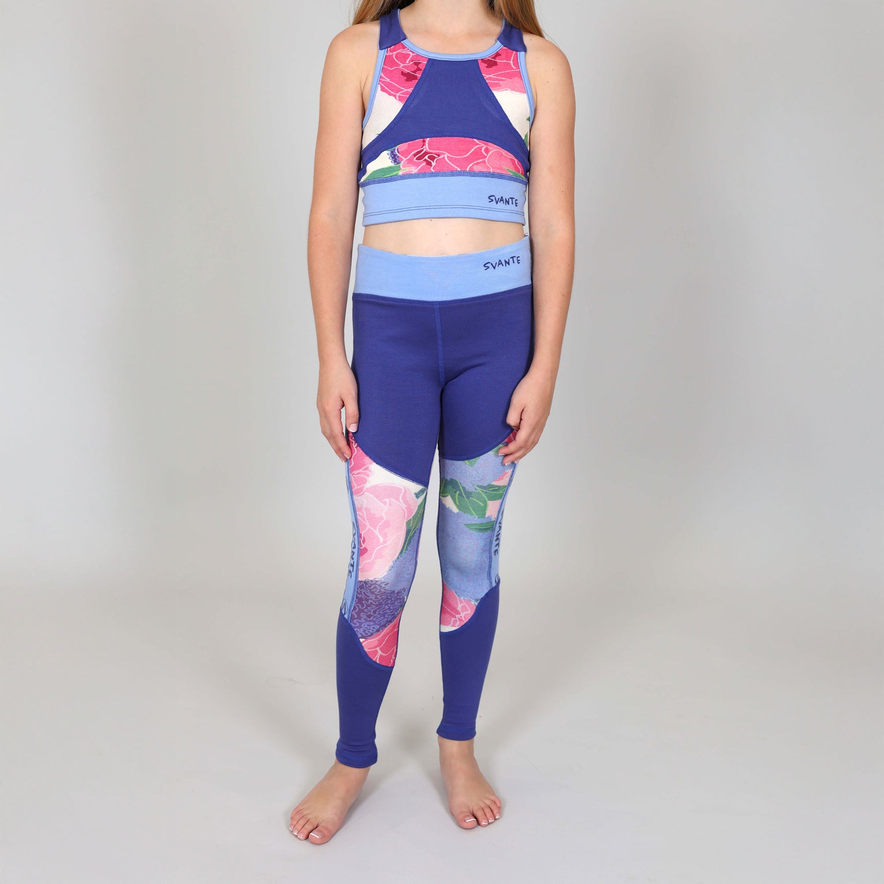 Girl wearing a colourful blue and floral sports crop-top and matching floral sports leggings set on a plain background. 