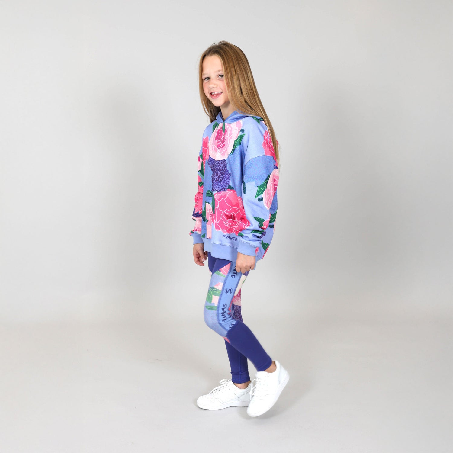 Girl aged eight standing side on wearing a floral organic cotton hoodie and matching sports leggings in a studio with grey background.  