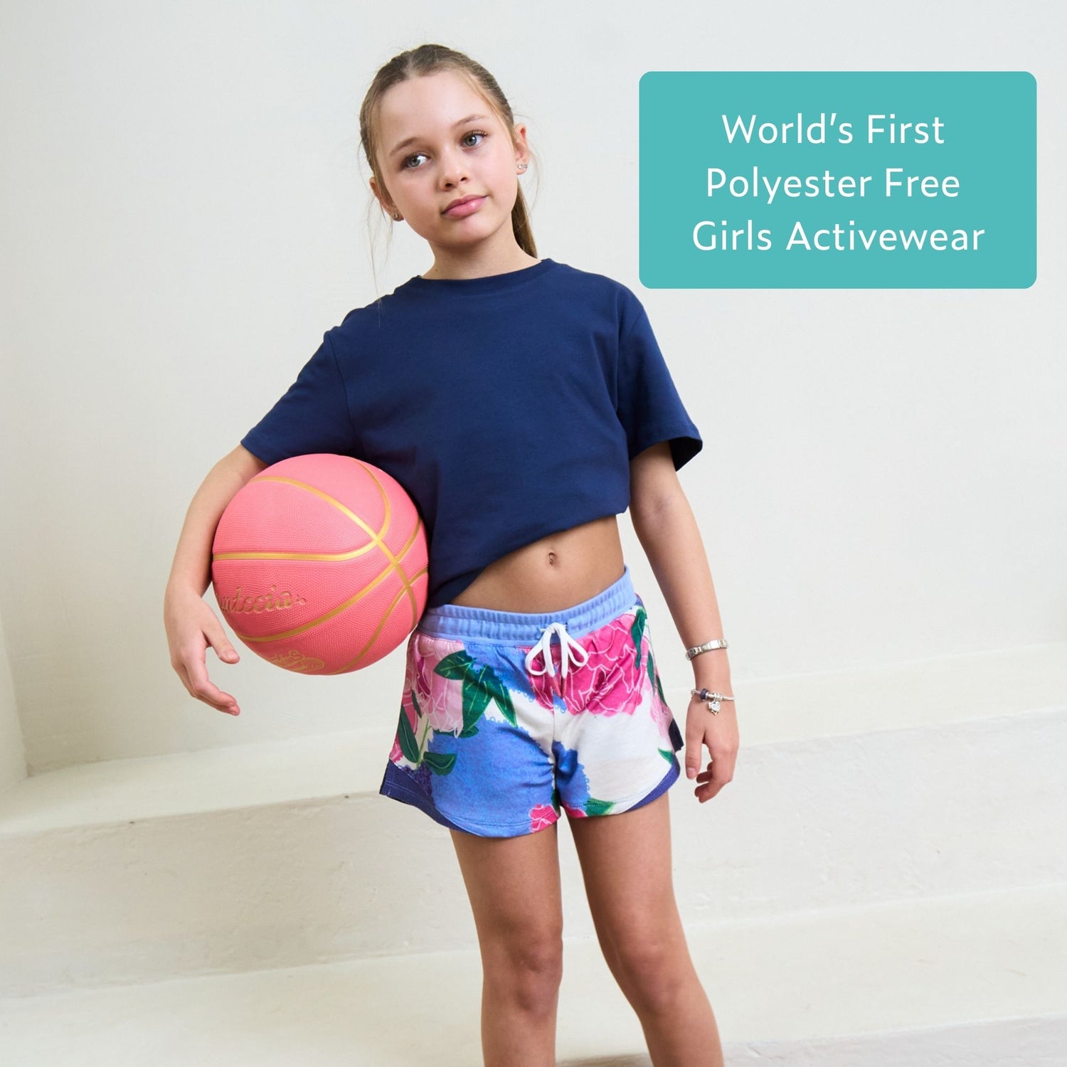 Girl holding a pink basketball wearing blue organic cotton t-shirt and blue sports shorts with a text overlay about polyester-free activewear on white background. 
