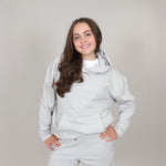 Female aged 12 with big smile and long hair, standing with hands on hips and wearing a grey sports organic hoodie and jogger on a plain background. 