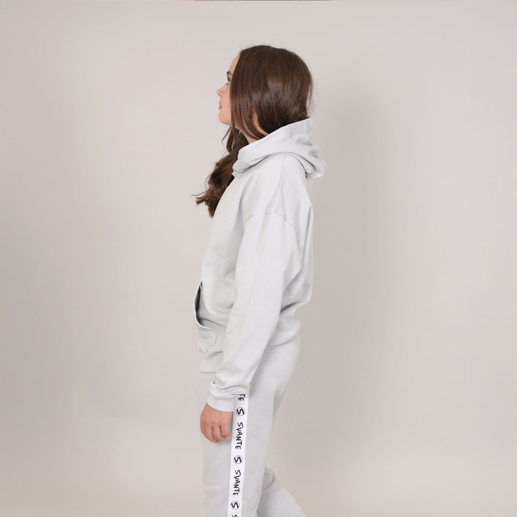 Girl aged 12 standing side on, looking upwards. She is showing the side view of a harbour mist (grey) hoodie and joggers with 'Svante' branding running down side of leg. 
