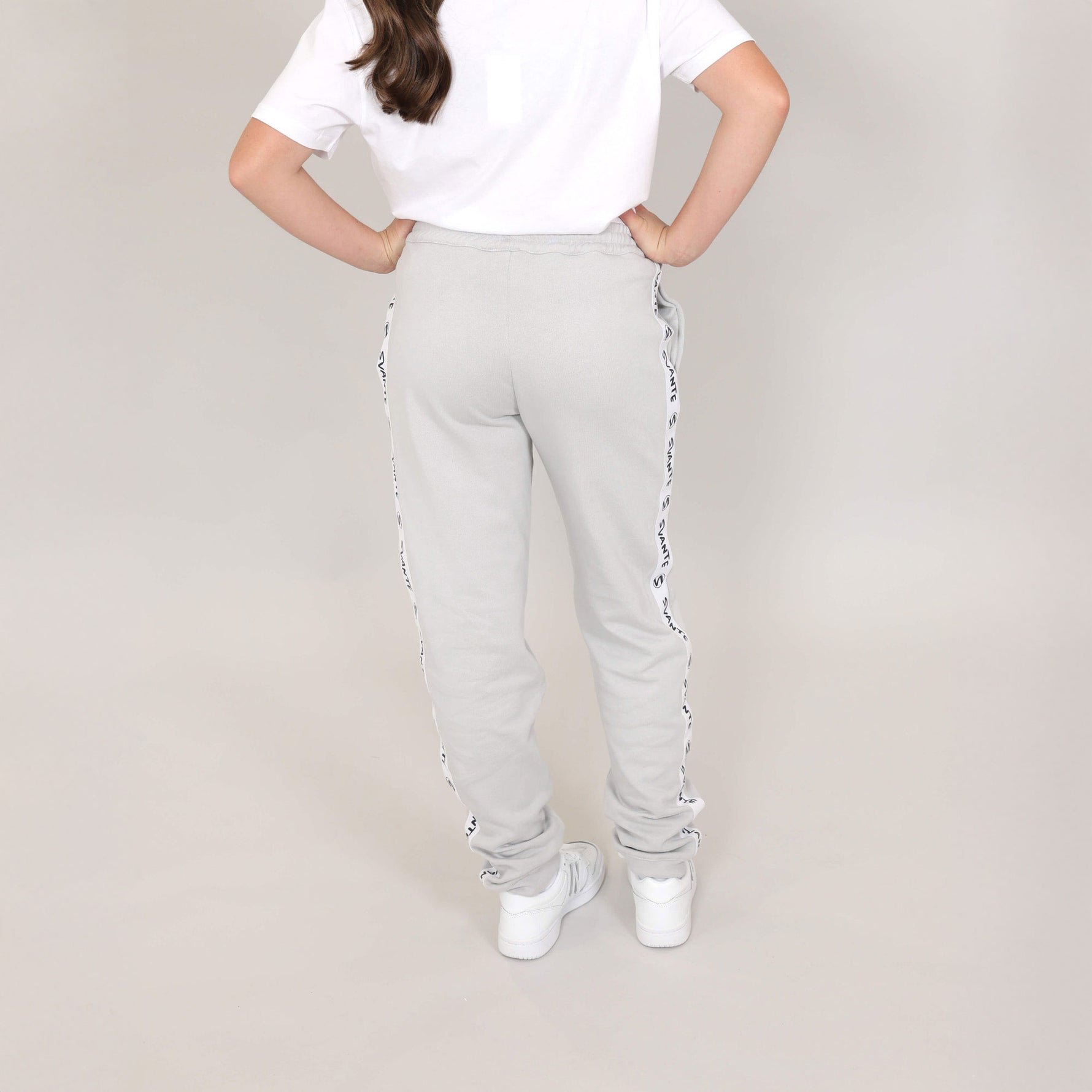 Teenage girl with back to photographer wearing light grey organic cotton sweatpants and white regenerative cotton t-shirt tucked in, with a logo pattern on the side, standing against a plain background. 