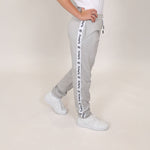 Girl with hands on hips showing organic cotton joggers with white and black tape 'Svante' branding down the leg and wearing white trainers on beige background. 