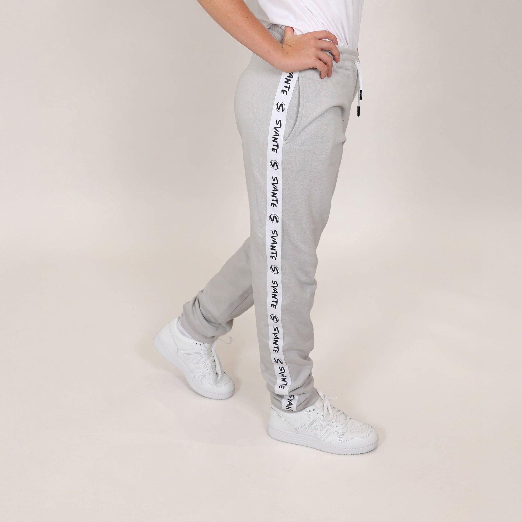Girl with hands on hips showing organic cotton joggers with white and black tape 'Svante' branding down the leg and wearing white trainers on beige background. 