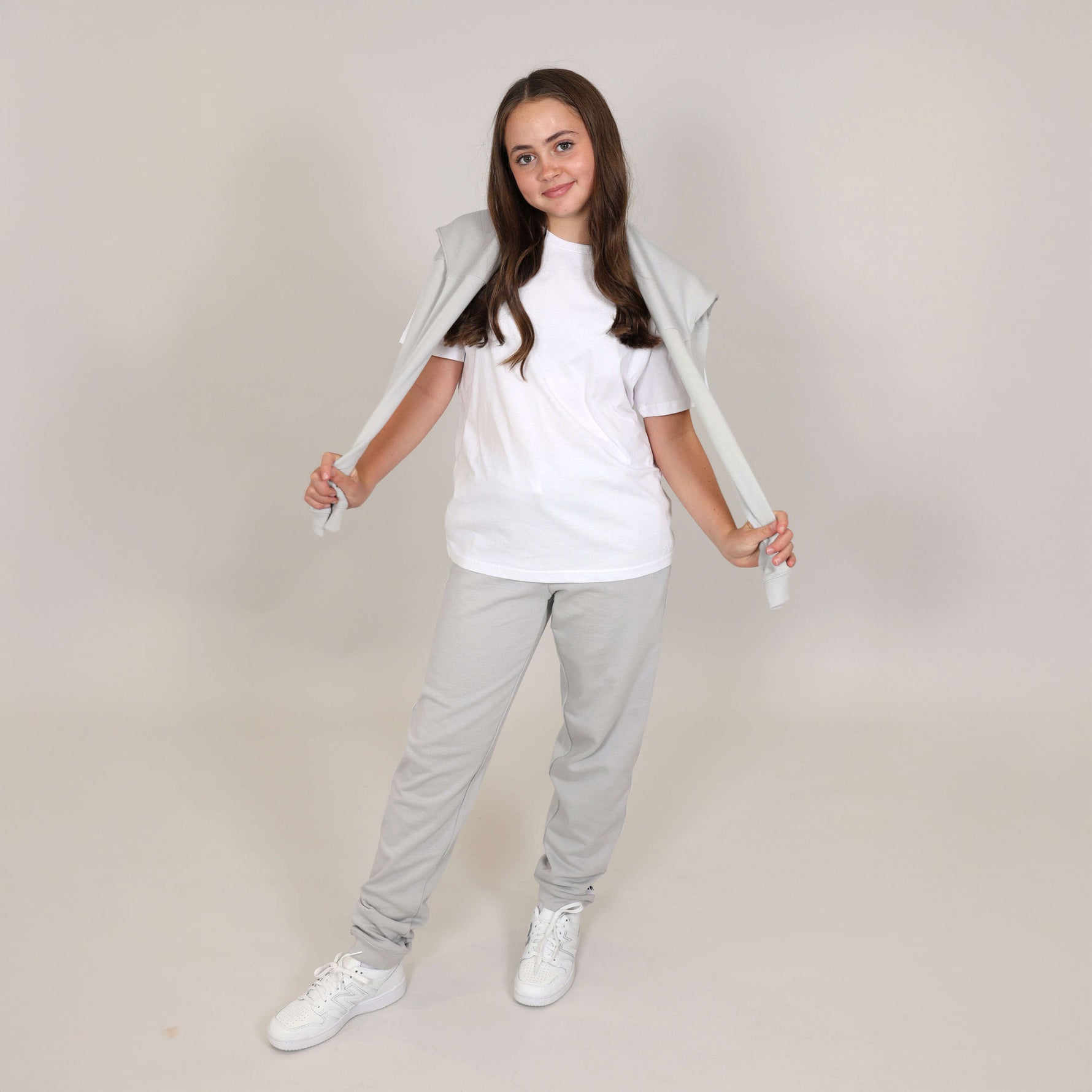 Teenage girl with nice smile wearing organic grey joggers, with regnerative cotton white t-shirt and white trainers, she has hoodie over her shoulders and pulling the arms out to the side. 