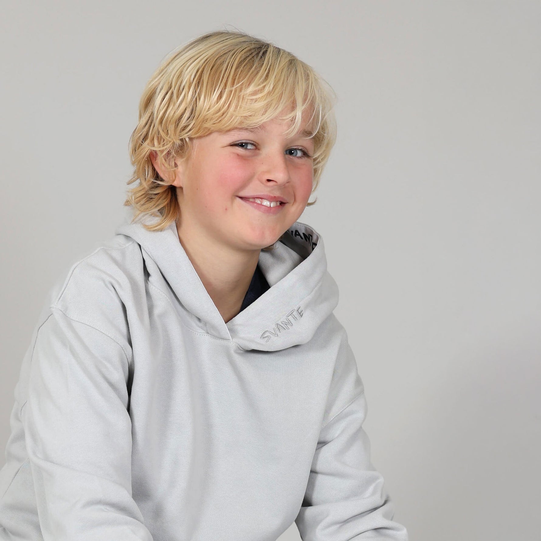 Boy smiling wearing a light grey organic cotton hoodie with 'Svante' logo a plain neutral background. 