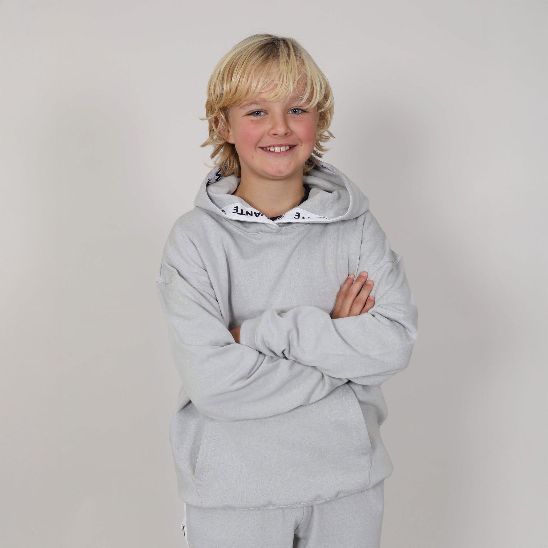Boy with arms folded wearing a grey organic hoodie with a white logo on a plain background. 