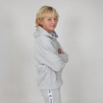 Boy with long blonde hair standing side on with arms folded wearing a light grey organic hoodie with a visible brand logo on a studio background. 