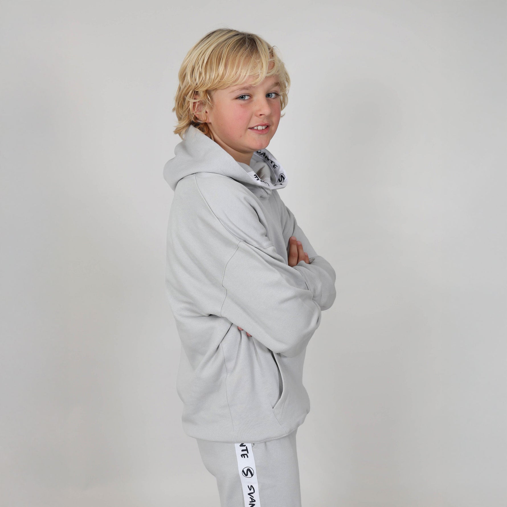 Boy with long blonde hair standing side on with arms folded wearing a light grey organic hoodie with a visible brand logo on a studio background. 