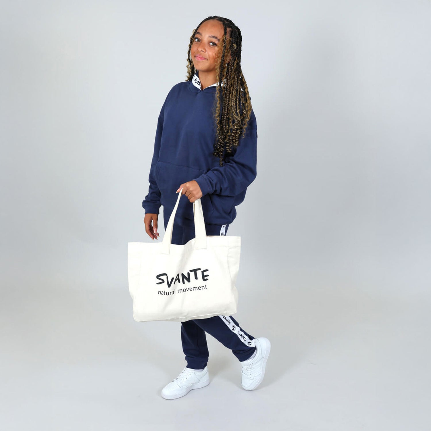 Teenage girl wearing organic navy blue hoodie and joggers, holding an organic cotton tote bag with 'SVANTE' branding on a plain background. 