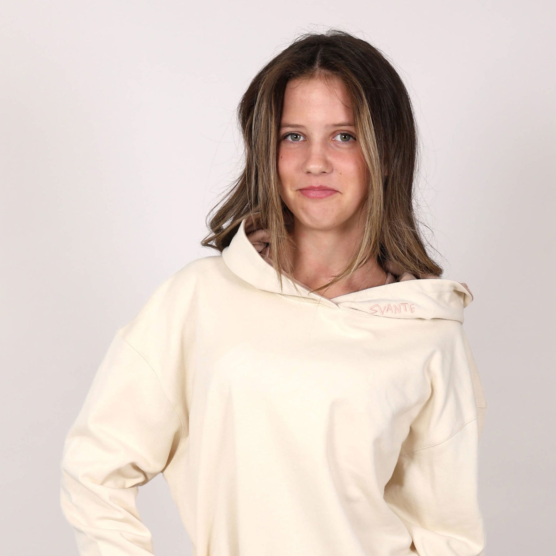 Close up front pictures of girl wearing a cream-coloured hoodie with 'Svante' branding on hood, against a pale grey background. 