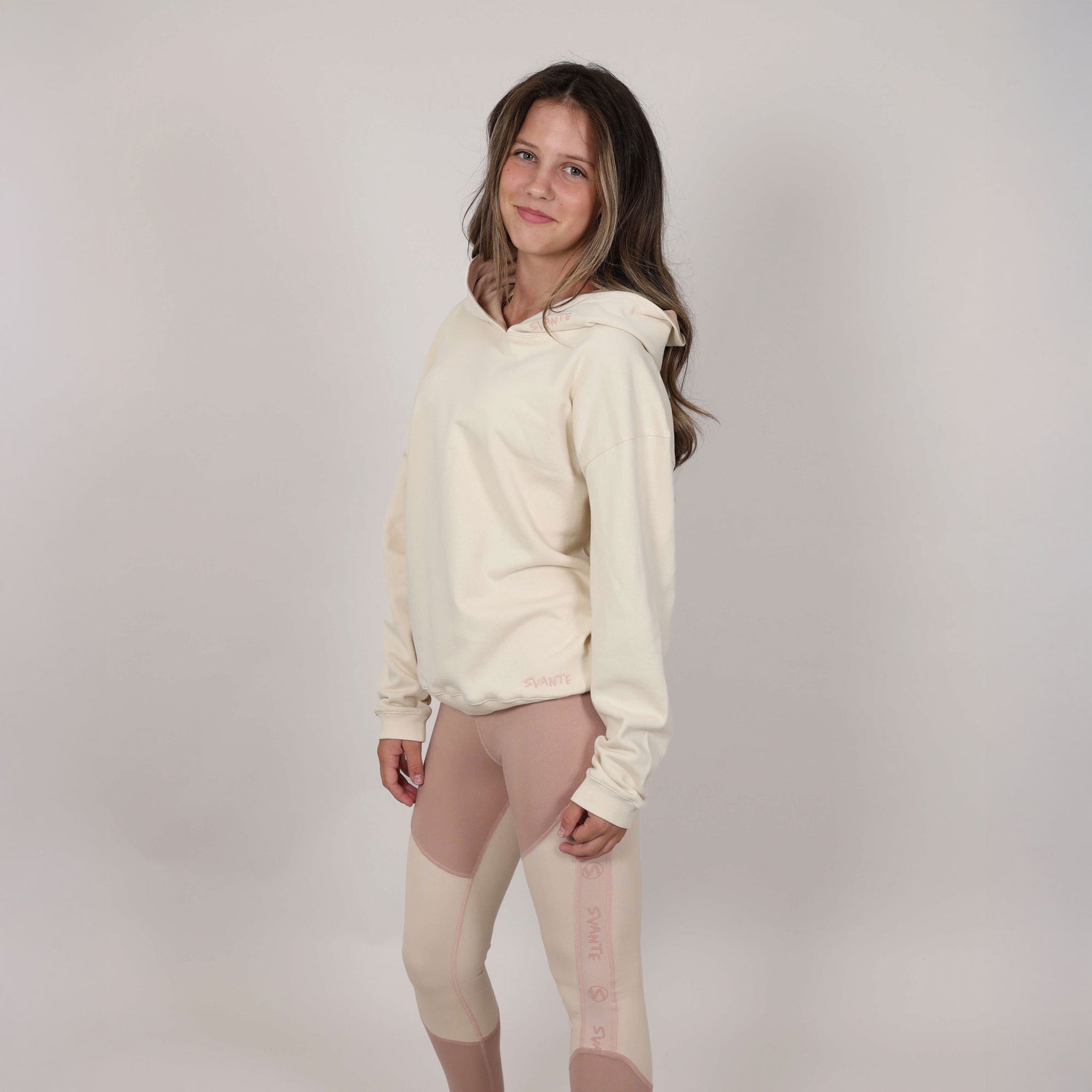 Teenage girl standing side on wearing a cream hoodie and leggings with Svante branding against a plain background. 